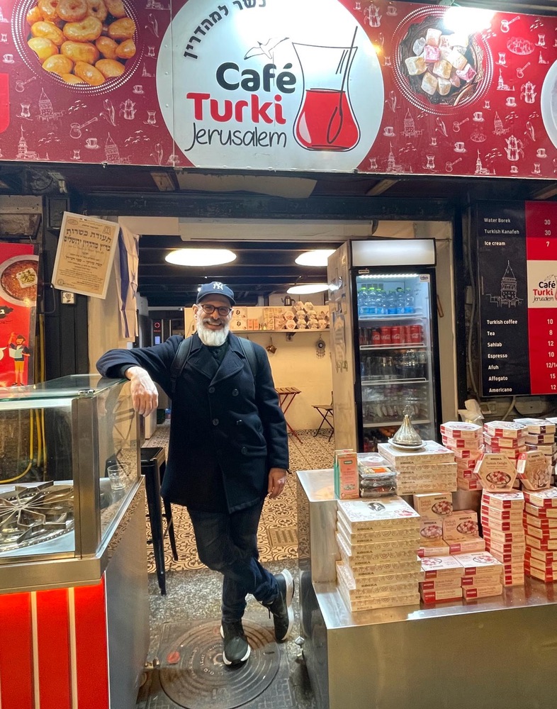 Harry Rubenstein standing in front of Cafe Turki in Mahane Yehuda Market with mehadrin kosher certificate visible above his shoulder.